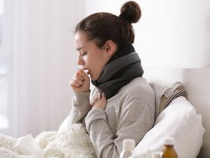 Eine Frau liegt krank im Bett und hustet.