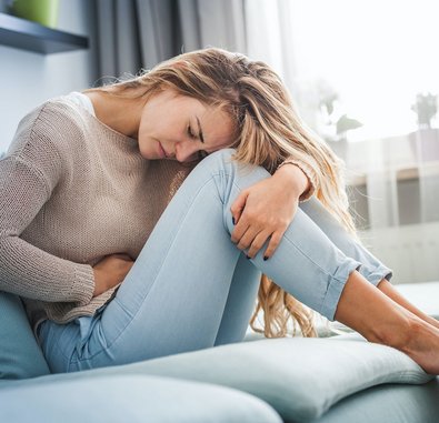 Eine Frau sitzt gekrümmt auf einer Couch und hält sich vor Schmerzen den Bauch